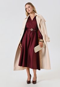 Abito a portafoglio borgogna con vita cinturata, abbinato a un trench beige, accessori dorati e una pochette scintillante. Scarpe con tacco completano il look.