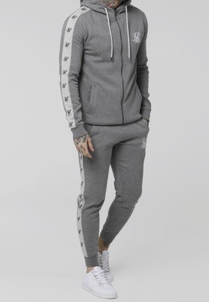 Sudadera con capucha gris con cremallera, cordones blancos y logo, combinada con pantalones jogger a juego con franjas laterales y detalles de logo. Zapatillas blancas.