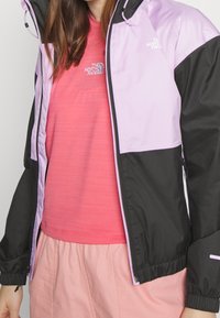 Lehká zkrácená bunda v černé a levandulové barvě s předním zipem, elastickými manžetami a logem na hrudi. Nositá přes růžovou košili.