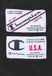 Two fabric labels on a black background. The top label is white with red accents and the text "Champion New York," the bottom is also white with "Champion U.S.A."