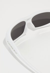 Λευκά γυαλιά ηλίου με σκούρους γκρι φακούς, αεροδυναμικό σχέδιο και γυαλιστερή φινίρισμα. Διαθέτουν λογότυπο στο πλάι και καμπυλωτά βραχίονες.