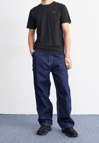 Černé tričko s krátkým rukávem a modrým logem, v kombinaci s volnými tmavě modrými džíny s bílým prošíváním. Doplňují černé tenisky.