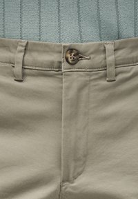 Κοντινή λήψη μπεζ παντελονιού με κούμπωμα με κουμπιά και θηλιές για ζώνη, φορεμένο με ανοιχτό μπλε πλεκτό τοπ με υφή, που έχει μπει μέσα.