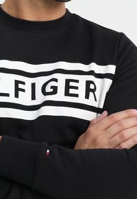 Sudadera negra con franjas horizontales blancas y el logo de "TOMMY HILFIGER". Presenta un cuello redondo y un pequeño acento de logo en la manga.