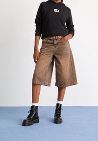 Pantaloni culotte in denim marrone con taglio dritto, indossati con una felpa nera con cappuccio e stivaletti neri con lacci. Calzini bianchi alla caviglia visibili.