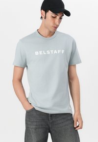 Világosszürke pamut póló, fehér "BELSTAFF" felirattal, rövid ujjakkal, kerek nyakkal, sötétszürke farmernadrággal párosítva.