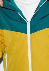 Bunte Jacke mit Reißverschluss, bestehend aus einem oberen Abschnitt in Türkis und einem unteren Abschnitt in Gelb. Hergestellt aus einem glatten, leichten Material mit weißem Reißverschluss.
