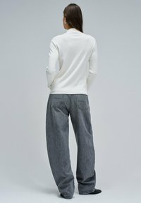 Vrouw met lang bruin haar in een witte blouse met lange mouwen, losse grijze jeans, zwarte schoenen, staande met handen in de zakken, met de rug naar de camera gericht op een grijze achtergrond.