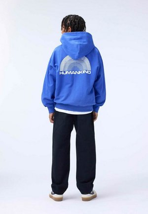 Felpa con cappuccio blu, dotata di coulisse, con una grafica bianca di linee orizzontali sopra al testo "HUMANKIND" sulla schiena. Pantaloni neri e sneakers.