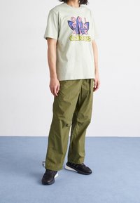 Světle zelené bavlněné tričko s nápisem "ORIGINALS" a modro-červeným logem. Olivově zelené lehké kalhoty. Černé tenisky s bílými detaily.