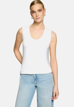 Femme blonde portant des boucles d'oreilles créoles dorées, un haut blanc sans manches à encolure dégagée et un jean bleu clair taille haute, debout devant un fond uni.