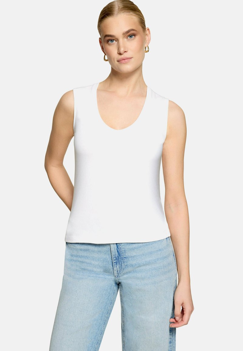 Femme blonde portant des boucles d'oreilles créoles dorées, un haut blanc sans manches à encolure dégagée et un jean bleu clair taille haute, debout devant un fond uni.