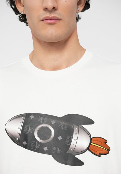 Κοντινό πλάνο ατόμου που φοράει λευκό πουκάμισο με μαύρο και ασημί γραφικό ρουκέτας και πορτοκαλί λεπτομέρεια φλόγας στο στήθος.