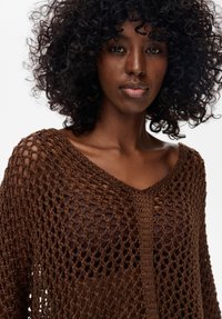 Brązowa, luźno dziergana bluzka z dekoltem w kształcie V, z teksturowanym wzorem. Lekki, półprzezroczysty materiał o otwartym splocie.