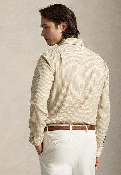 Jeune homme aux cheveux foncés tourné de profil, portant une chemise beige à rayures rentrée dans un pantalon blanc avec une ceinture marron, main dans la poche.