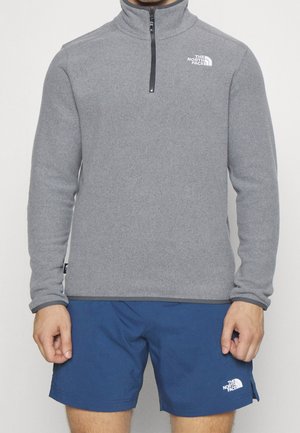 Mann trägt ein graues Fleecepullover mit Viertel-Reißverschluss und blaue Shorts, beide mit weißen The North Face-Logos versehen, steht mit entspannten Händen.