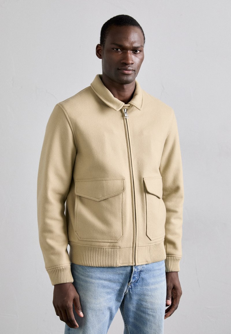 Beige wolmix jas met een kraag, ritssluiting aan de voorkant en twee grote opgestikte zakken. Geribde cuffs en zoom, gecombineerd met lichtblauwe jeans.