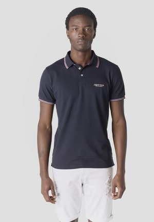 Joven con una camiseta tipo polo azul marino con detalles en rojo y blanco, y pantalones cortos blancos desgastados, de pie sobre un fondo claro y liso.