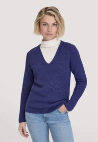 V-Ausschnitt-Pullover in tiefblau, mit gerippter Struktur, langen Ärmeln und legerem Schnitt, getragen über einem weißen Rollkragenpullover. Einfaches und modernes Design.