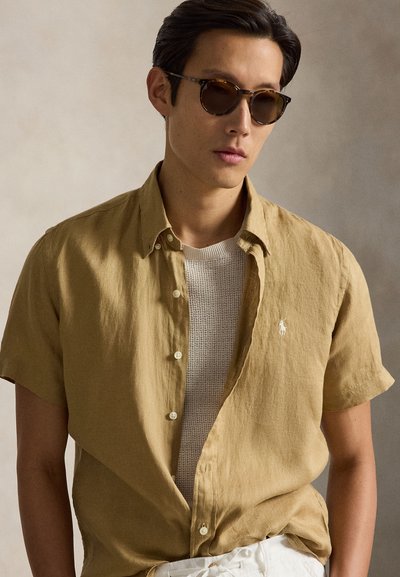 Άνδρας που φοράει γυαλιά ηλίου με σκελετό από χελώνα, μπεζ κοντομάνικο πουκάμισο με κουμπιά πάνω από λευκό πλεκτό τοπ, στέκεται μπροστά σε ουδέτερο φόντο.