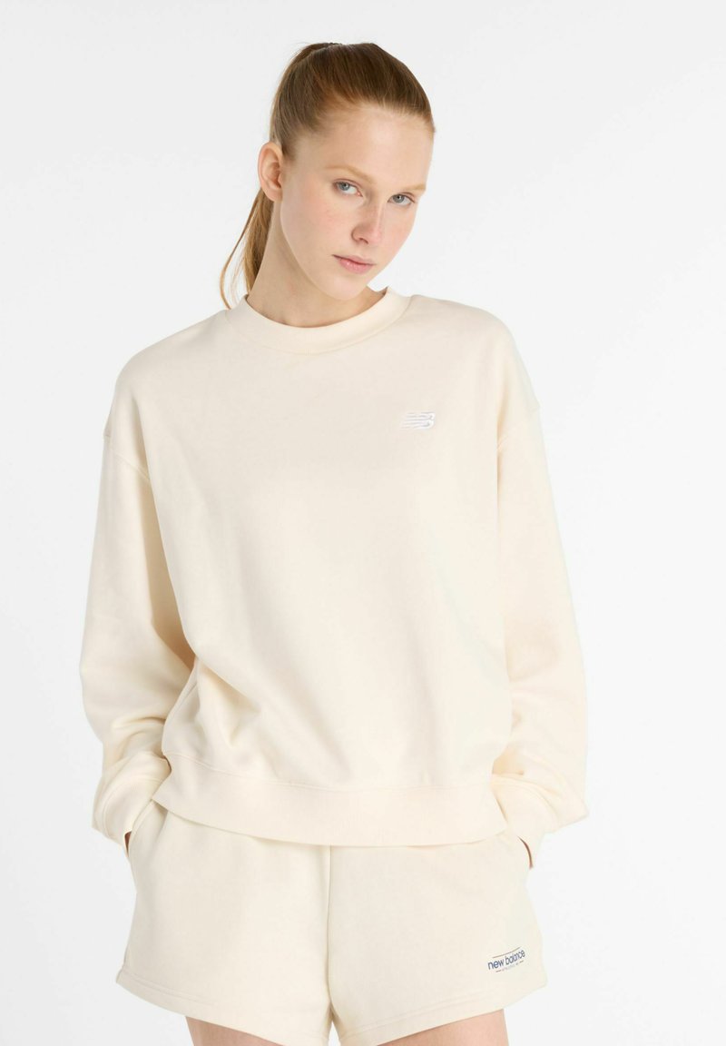 Sudadera color crema hecha de tejido suave, con cuello redondo y un pequeño logo blanco en el pecho. Presenta un corte relajado y puños de canalé.
