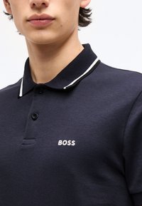 Νεαρό άτομο που φοράει μπλε σκούρο πόλο μπλούζα με μαύρο γιακά και λευκή διακόσμηση, με τη λέξη «BOSS» κεντημένη στο στήθος.