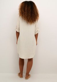 Abito in lino beige chiaro con maniche arrotolate, vestibilità ampia e lunghezza fino a metà coscia. Tessuto semplice e strutturato con un design minimalista.