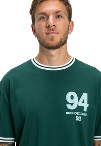 Młody mężczyzna z jasną brodą, ubrany w ciemnozieloną koszulkę z białymi paskami przy kołnierzyku i rękawach, z nadrukiem "94 DCSHOECOUSA" na piersi.