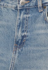 Jean en denim bleu en gros plan montrant une poche avant, des coutures orange, une boucle de ceinture et un bouton en métal avec gravure de la marque.