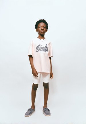 Junge, der ein hellrosa grafisches T-Shirt, weiße Shorts und graue Freizeitschuhe vor einem einfarbigen weißen Hintergrund trägt.