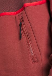 Rote Kapuzenjacke mit einem strukturierten, dunkelbraunen Panel oben. Verfügt über eine Reißverschlusstasche mit schwarzem Reißverschluss und Steppdetails.