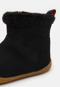 Botín negro de ante con forro de piel sintética. Presenta una suela de goma marrón y una pestaña de tirón con un acento rojo. Textura suave y diseño simple.