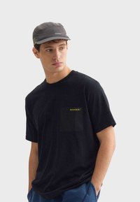 Černé tričko s krátkým rukávem a kapsou, na kterém je žluté logo. Šedá kšiltovka s texturovaným povrchem a kontrastními zelenými akcenty.