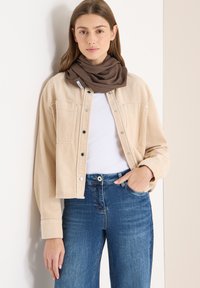 Veste en velours côtelé beige avec boutons-pression, portée avec un haut blanc et un jean bleu. Écharpe marron bouclée autour du cou.