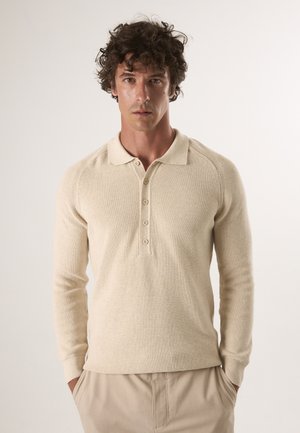 Beige gebreide pullover met een polo-kraag, vijfknopenlijst, ribbelstructuur en aansluitende lange mouwen. Draagt met lichtgekleurde broeken.