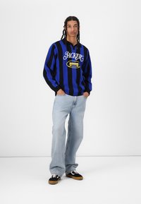 Blå og svart stripete genser med 'JACKER' logo, kombinert med lyseblå bredbenede jeans og svarte sneakers med brune detaljer.