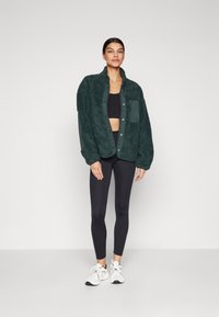Grüne Fleece-Shirtjacke mit strukturiertem Finish, ausgestattet mit Knöpfen, einer Brusttasche und einer lockeren Passform, kombiniert mit schwarzen Leggings und weißen Sneakers.