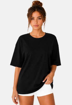 Jonge vrouw met bruin haar in een knot, draagt een los zwart T-shirt en witte korte broek, staand tegen een effen lichte achtergrond.