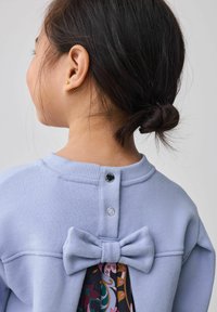 Ragazza giovane con capelli scuri raccolti in un chignon, che indossa un maglione blu chiaro con dettagli di bottoni sul retro e un grande fiocco di tessuto sopra un abbigliamento a fiori.