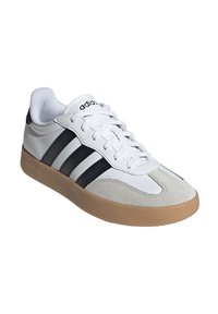 Baskets Adidas avec une tige en cuir blanc, une pointe en daim gris, des bandes noires et une semelle en caoutchouc gomme. Les lacets sont blancs.