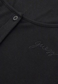 Black ribbed top featuring a round neckline, a button closure, and embroidered "Guess" logo in cursive on the front. Textured fabric detail.