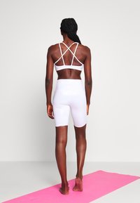 White sports bra with crisscross straps, paired with white high-waisted shorts. Smooth texture, minimalistic design, displayed against a pink mat.