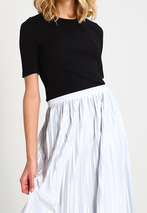 Vrouw draagt een zwart topje met korte mouwen en een witte plissérok met hoge taille, houdt de rok met één hand vast.