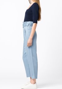 Femme aux longs cheveux portant un haut bleu marine ajusté, un pantalon tailleur bleu clair taille haute avec une ceinture, et des baskets blanches, debout de profil.