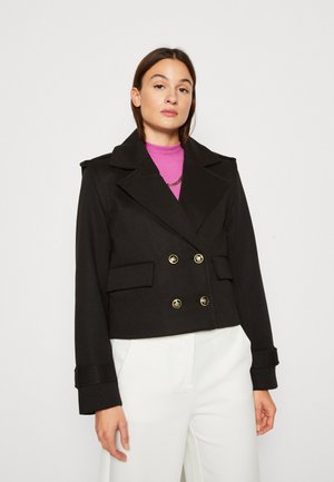 Veste noire courte avec fermeture à double bouton doré, large col et deux poches avant. Associée à un col roulé rose.
