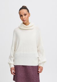 Pull à col montant en tricot crème doux avec de larges manches ; associé à une jupe en feutre bordeaux. Présente des poignets en côte et une silhouette droite.