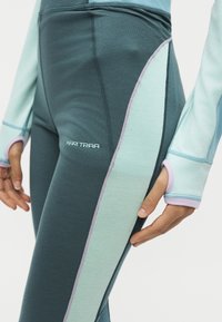 Gros plan d'une personne portant un legging bleu et turquoise avec le logo Kari Traa et un haut à manches longues assorti avec des trous pour les pouces au niveau des manches.