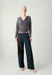Gray draped long-sleeve top paired with wide-leg dark pants. Model holds a light-colored bag, wearing open-toe sandals on a plain background.