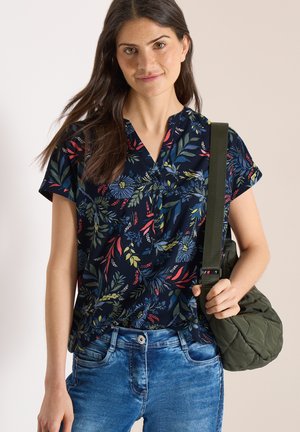 Młoda kobieta o długich ciemnych włosach, ubrana w granatową, kwiatową bluzkę z krótkim rękawem, niebieskie dżinsy, niosąca zieloną pikowaną torbę na ramię.