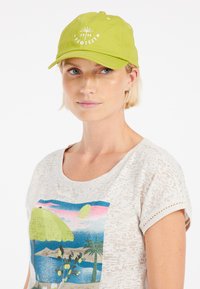 Groene honkbalpet met een gebogen rand en witte geborduurde tekst met een zonnestraalontwerp. De tekst bevat de merknaam "PROTEST."